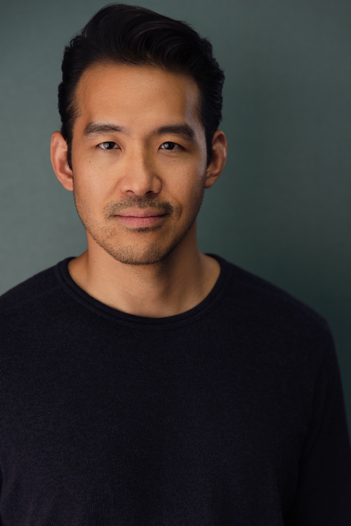 Alex Chung, a voice actor with swept-back dark hair, looking directly at the camera against a muted green backdrop.